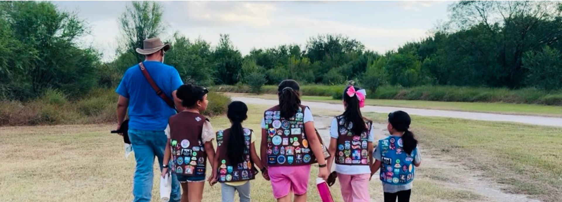  adult volunteer wearing vest with a girl scout junior (right, in baseball cap) and daisy (left) outside at a park smiling and looking at one another 