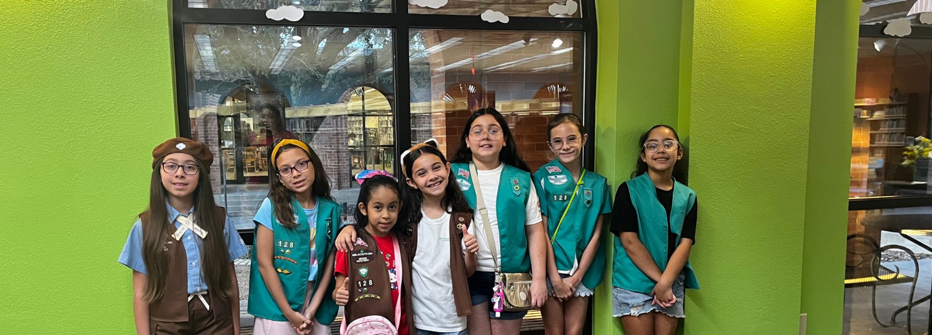  two girl scouts wearing trefoil clothing leaning on one another indoors 